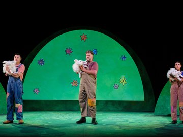 Three actors dressed in farmer overalls on a stage with a cartoonish green backdrop, each holding a fluffy white sheep puppet