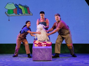 Three actors in overalls and t-shirts surround a large white stuffed sheep on a bed set-piece, with a painted cartoon background of a bookshelf and lamp.
