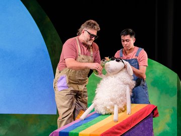 Two actors in overalls and t-shirts, one pouring liquid for a fluffy white sheep puppet wearing sunglasses, lying on a rainbow-striped towel, on stage with blue and green painted backdrop.