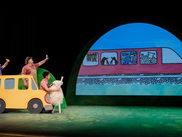 A theatrical production featuring three actors in front of a yellow car prop, waving to a projected image of a cartoon train with various animals, including fish, penguins, and a sheep, inside.