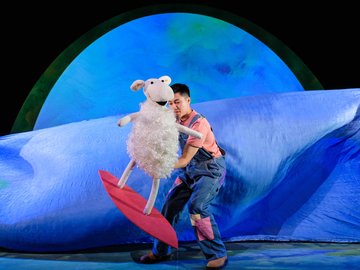 A man dressed in overalls holds a fluffy sheep puppet on a red surfboard, performing on stage with a blue backdrop simulating water.