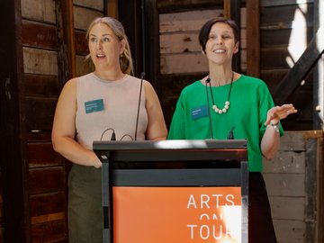 Two women speaking at a podium with "ARTS ON TOUR" displayed, one in a green top gesturing with her hands.