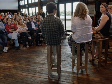 A panel discussion with four people seated on high wooden stools, facing an audience seated on chairs in a large open space with wooden floors and expansive windows.