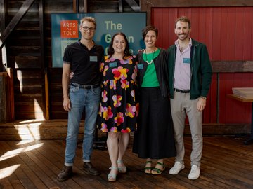 Four people standing together and smiling in front of a screen displaying the words "Arts on Tour" and "The Green Circle". They are dressed in smart casual attire. The background has wooden and red walls.