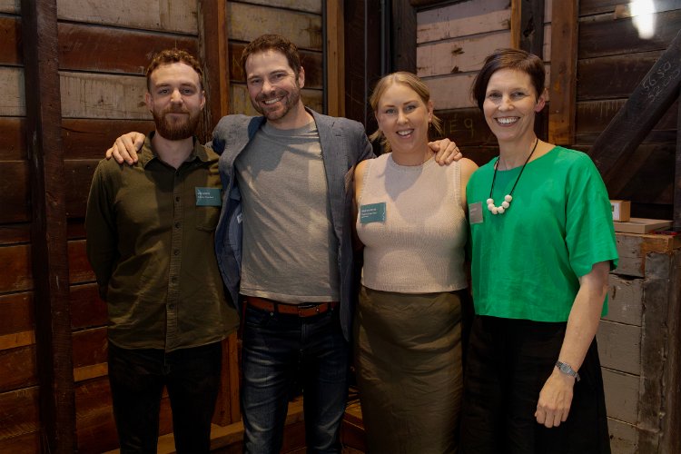 Four people, two men and two women, pose for a photo with their arms around each other's shoulders, smiling at the camera, wearing name tags, and standing in front of a wood paneled wall.