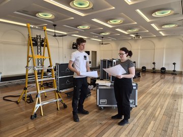 Two individuals review plans near a large yellow ladder and stacked equipment cases in a spacious room with a wooden floor and ornate ceiling lights.