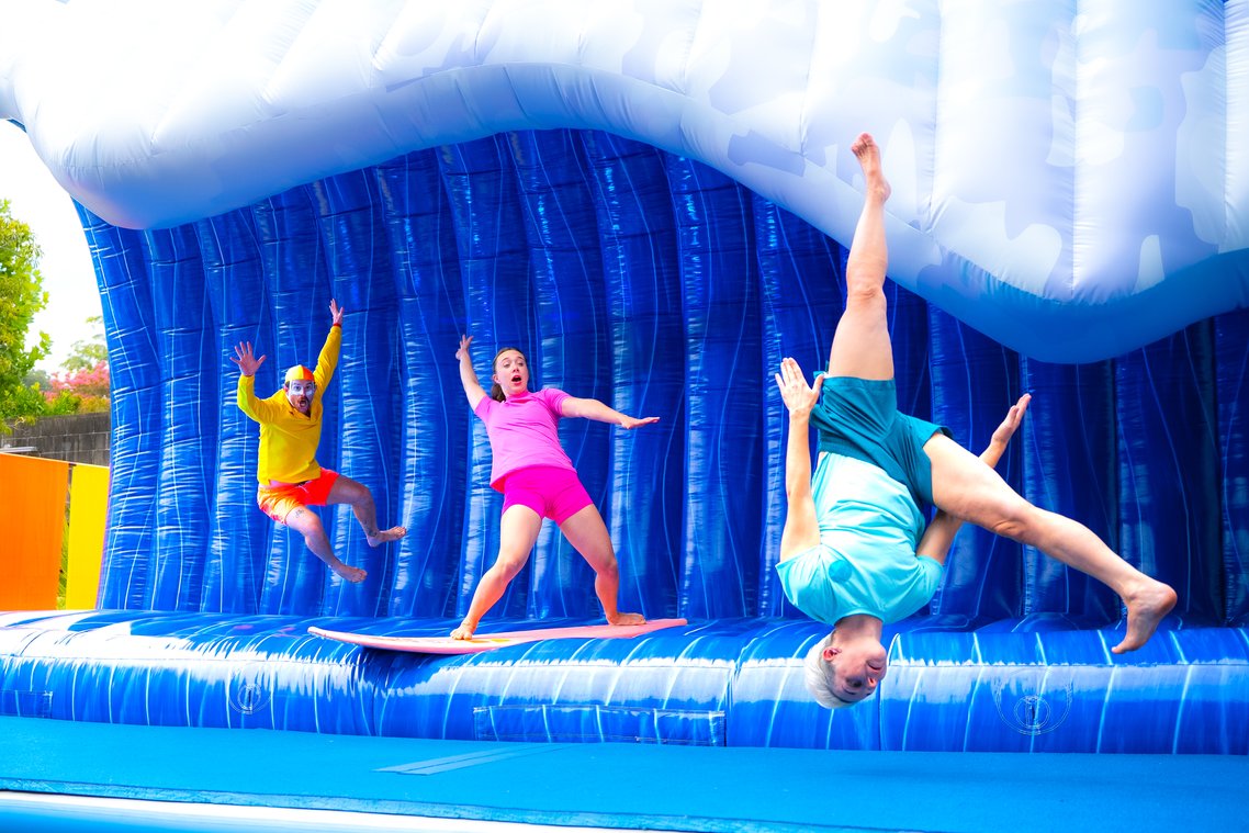 Three physical theatre performers pretending to surf an inflatable blue wave. One person wears surf life saver attire, another stands on a surfboard, wearing pink swimwear. The third person is upside down, with legs in the air, wearing blue swimwear.