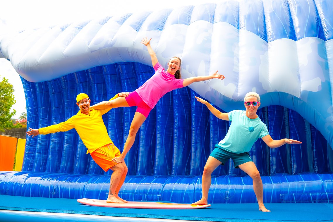 Three individuals in bright, contrasting swimwear strike playful poses on a surfboard against a large, blue, inflatable wave structure.