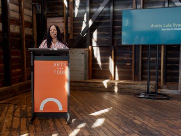 A woman with long black hair, wearing a floral-patterned shirt, speaking at a podium with "Arts on Tour" branding in an old wooden building, and a screen in the background displaying "Aunty Lola Ryan: Welcome to Country".