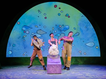 Three actors in costume performing on a stage with a cartoon-style backdrop depicting bubbles; a stuffed sheep is in a bathrub on a purple pedestal.