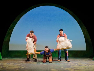 Three actors on a stage with a whimsical backdrop, two holding large plush sheep on either side of a wooden beam, and a third actor on their knees balancing on the beam.