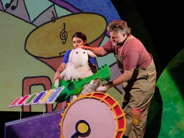 Two people arranging a plush sheep playing a green guitar on a stage in front of a colourful backdrop, with a xylophone and drum below.