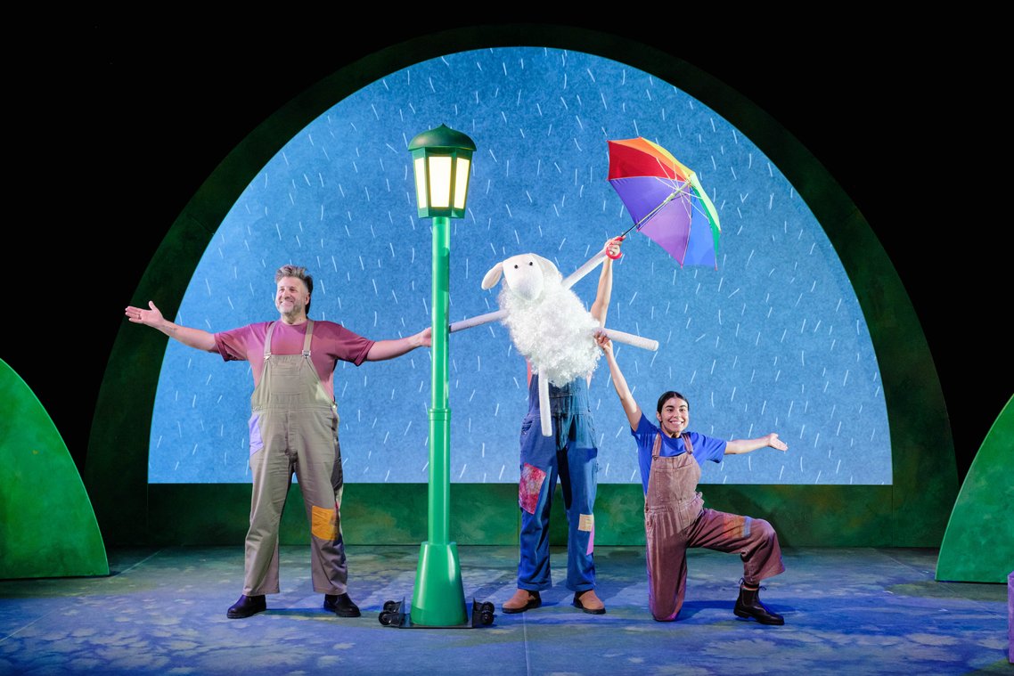 Three actors in colourful overalls on stage, one holding a sheep puppet and rainbow umbrella, another a green street lamp, all posing with arms outstretched, against a backdrop of a blue sky with rain.