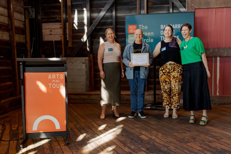 Four people stand on a wooden stage, smiling, with one person holding a certificate. A lectern with the words "ARTS ON TOUR" is on the left; a backdrop with logos of "ARTS ON TOUR" and "The Green Circle" is on the right.