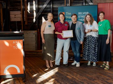 Five people standing on a wooden floor, holding a certificate and small gifts, in front of a teal banner with white text in a wood-paneled room. One woman in a red shirt and jeans holds a framed certificate. A podium with "ARTS TOUR" printed on an orange 