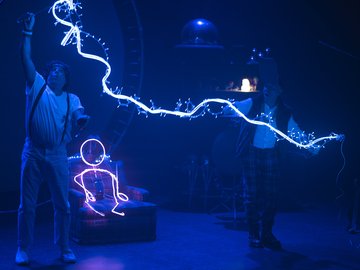Two men perform with strings of white lights in a dimly lit blue-lit space; one man holds the lights above his head, creating a zig-zag pattern, while the other man holds the end of the light string and a stick figure drawn with light sits in an armchair.