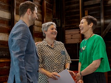 Three people laughing together in a wooden-paneled room: a man in a blue blazer, a woman in a black and white patterned dress holding a paper, and a woman in a green top with a necklace.