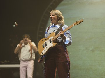 A female musician with blonde hair sings and plays a light-brown electric guitar on a stage, wearing a blue and white patterned shirt, brown pants with small lights along the seams, and a black guitar strap.