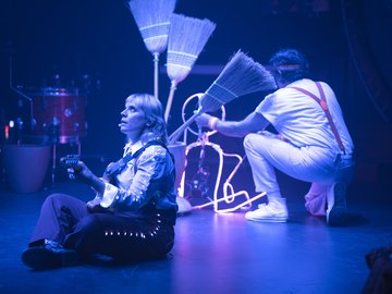 A woman with blonde hair sits cross-legged on a stage, playing an electric guitar under blue stage lights. A man wearing white pants, shirt, and suspenders crouches nearby, holding a broom towards a glowing neon outline of a figure.