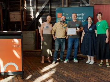 Two men and three women standing together, posing with an award, in an old wood-paneled building for an Arts on Tour event. A podium with "ARTS ON TOUR" is visible in the foreground. In the background a sign for "The Green Circle".