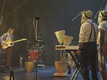 A quirky band performing on stage. A guitarist in a sequined jacket plays a yellow electric guitar. Two band members, wearing insect-like masks and suspenders, stand by a keyboard with attached brooms. A drum kit is also present.