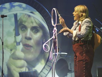Female musician in vintage attire, performing onstage with a light sculpture in front of her, a large projection of her face in a circular frame behind her.