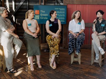 Five people sit on wooden stools on a rustic wooden floor, participating in a panel discussion titled "The Green Circle". The person on the right is speaking into a microphone, and the others sit listening.