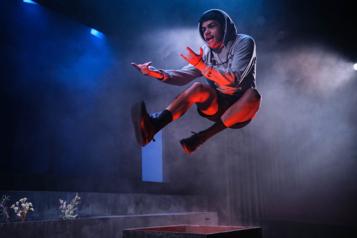 A young man in a grey Champion hoodie and black shorts, mid-air in an acrobatic pose, arms outstretched, hands gesturing "rock on", mouth open, lit by red and blue stage lighting with a foggy background.