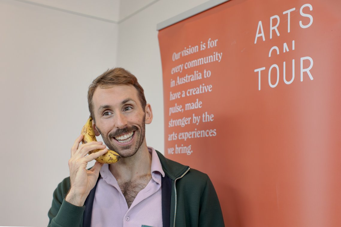 Man smiling and holding a banana to his ear like a phone in front of a banner that says "ARTS ON TOUR".