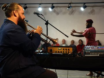 A profile shot of a bearded man playing a trumpet. He is sitting in front of a sound desk. Another man is dancing in the background with people watching.