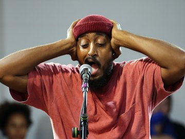 A mid-shot of a man at a microphone. He is wearing a maroon beanie and red t-shirt and has his hands on his head.