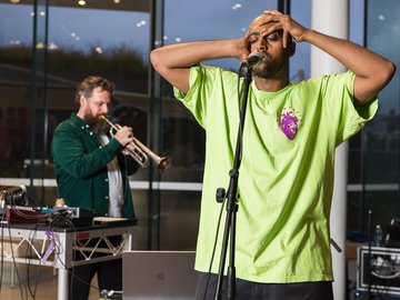 A mid-shot of a man at a microphone. He is wearing a lime green t-shirt and has his hands on his head. A bearded man plays the trumpet behind him.