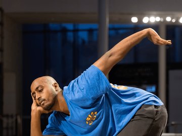 A full body shot of a man dancing. He is wearing a blue t-shirt and black pants. He us striking a pose with his right hand on his face and left hand outstretched. 