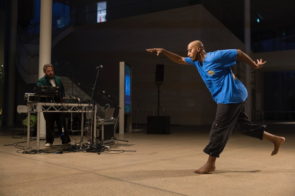 A full body shot of a man dancing. He is wearing a blue t-shirt and black pants. Another man is standing in the background with a sound desk.