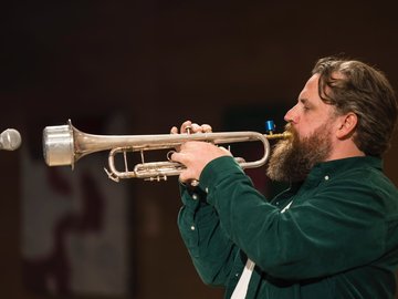 A profile shot of a bearded man playing a trumpet into a microphone. He is wearing a dark green, long-sleeve shirt.
