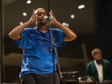 A mid-shot of a man at a microphone. He is wearing a blue t-shirt and has his hands raised near the microphone.