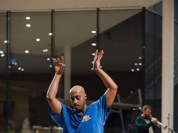 A full body shot of a man dancing. He is wearing a blue t-shirt and loose black pants.