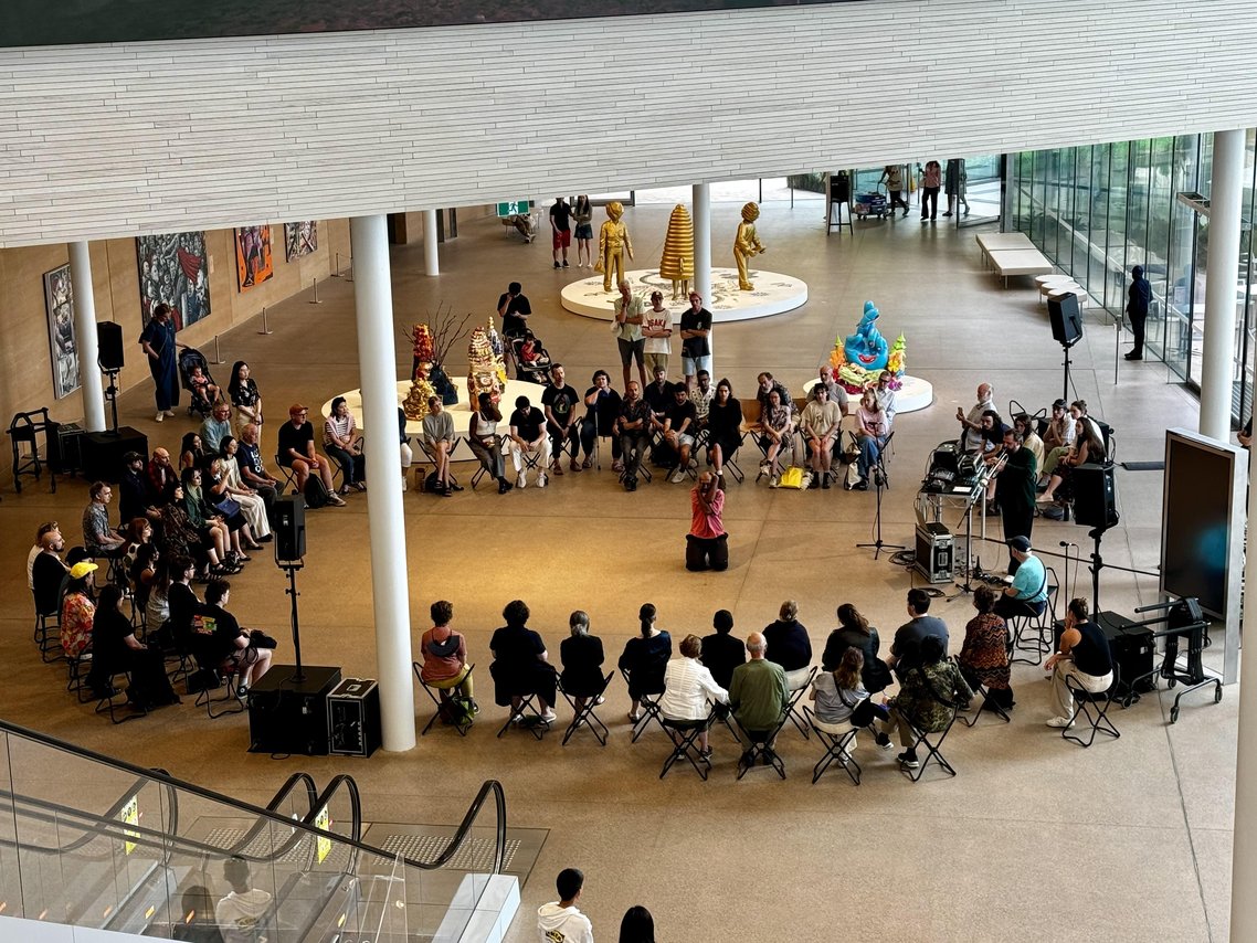 An aerial view of a large group of people sitting on chairs in a circle. There is a musician and dancer performing for the in the centre.