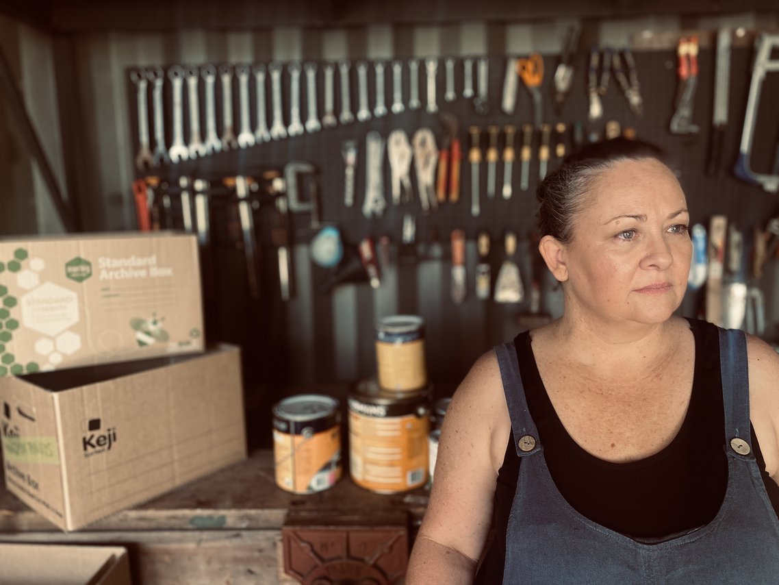 A woman in blue denim overalls and a black shirt stands in a workshop, looking off to the side, with a wall of tools behind her; boxes and paint cans sit on a shelf in the foreground.