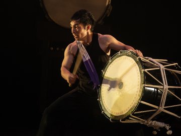 A man with dark hair, wearing a black tank top and pants, playing a large taiko drum with drumsticks, illuminated against a dark background. He has a purple and white sash around his neck and is mid-strike.