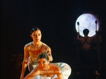 Two female dancers in neutral tones perform a choreographed routine on a dark stage, one kneeling and the other standing behind her, with a large, illuminated drum being played in the background.