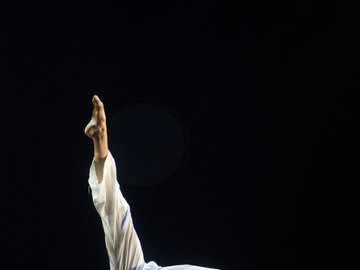 Female dancer in white flowing trousers and a light-colored top, performing a curved pose on a blue-toned stage with one leg extended upwards and head bent towards the floor.