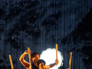 Two male performers in black attire perform Taiko drumming, holding wooden drumsticks in front of a large white Taiko drum, with a dark, abstract backdrop.