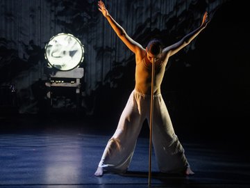 A ballet dancer in white pants and a flesh-toned leotard balances a long golden rod on their nose, arms outstretched, performing on a dark stage with a large, lit circular screen behind them.