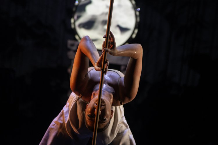 A female dancer in a white outfit, bending backwards and gripping a pole, with a projected image in the background.
