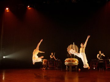 Two female performers execute mid-air leaps over Taiko drums on a stage, lit with spotlights, during a theatrical performance. Two other drummers are positioned further back, seated, and hitting their drums.