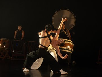 A female dancer in black pants and white top and male drummer in black attire perform a fluid movement in front of a giant taiko drum. Another drummer and several other taiko drums are visible on a dimly lit stage.