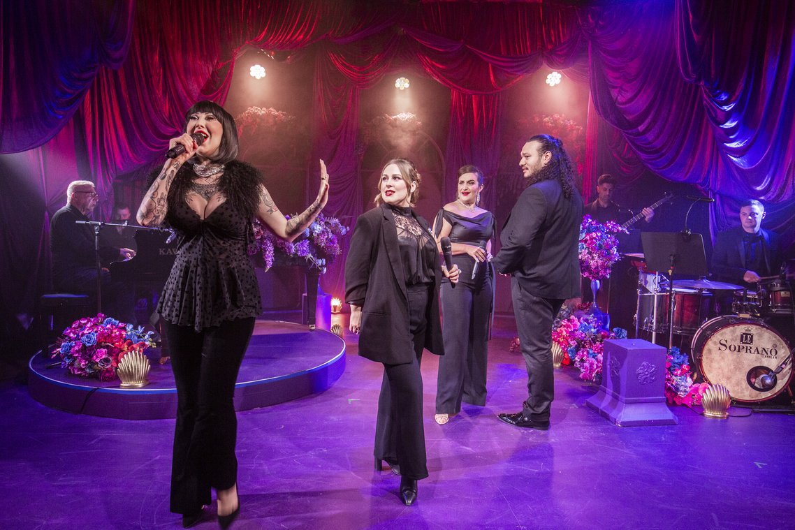 Four singers on stage, dressed in black. A band is behind them on a stage with purple theatrical lighting.