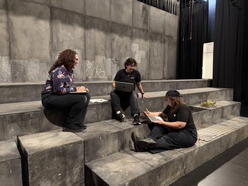 Three individuals sitting on concrete steps on a stage, two of them using laptops.