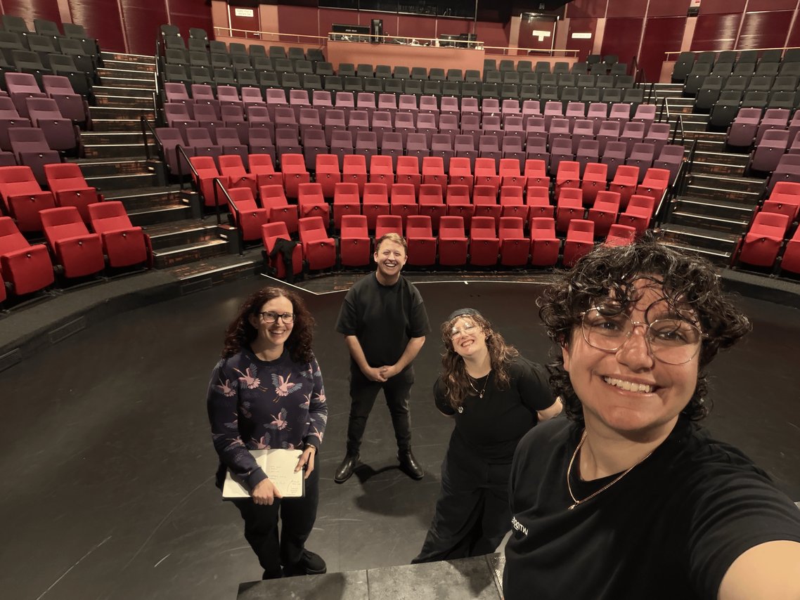 Four people stand on a stage in an empty theater with tiered seating in the background.